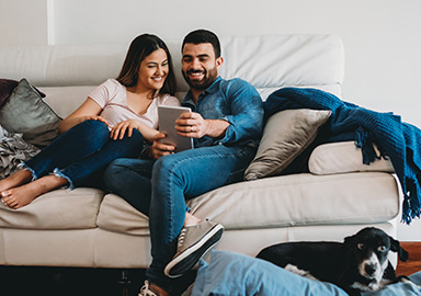 Couple sitting on beige sofa. Black dog with white markings lying on the floor.