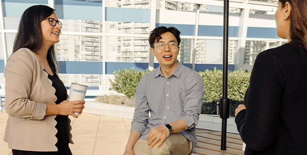 A woman wearing a beige blazer holding a travel mug, a man wearing glasses and a light blue shirt and a woman wearing a black blazer chatting on a roof top. 