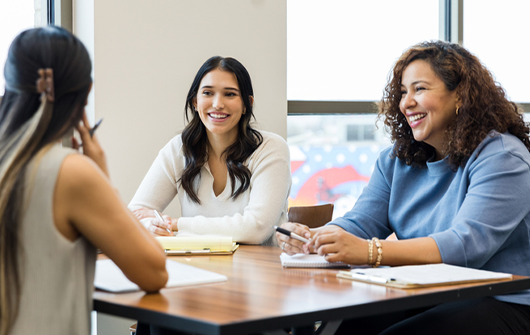 Three women sitting at table.  Two women are facing camera and the one woman has her back to the camera.