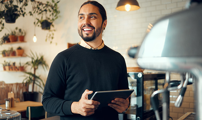 Smiling bearded man wearing blue crew neck sweater holding a tablet device.  