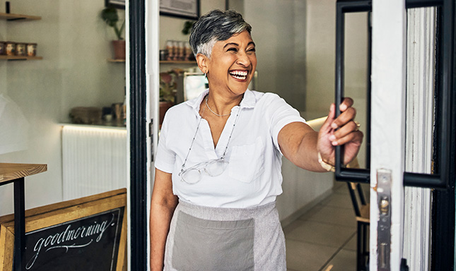 Smiling grey haired lady wearing a white shirt and grey apron standing in doorway holding the door open.