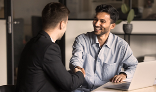 Two people shaking hands across a desk beside a laptop during a business meeting.