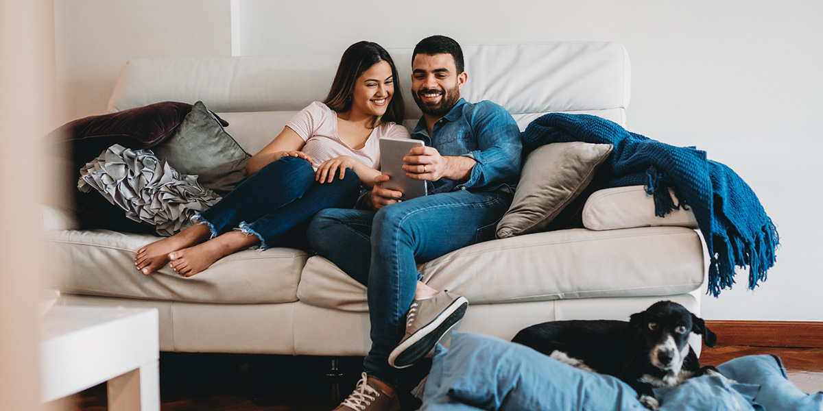 Couple sitting on beige sofa. Black dog with white markings lying on the floor.