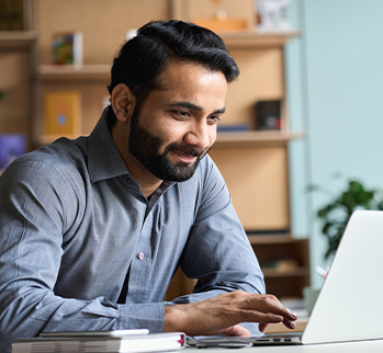 A bearded man with a grey shirt is sitting looking at a laptop