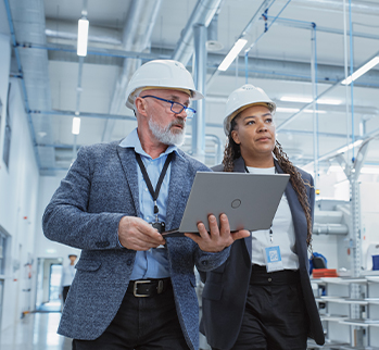 Woman and man wearing hard hats in a factory, holding a laptop.