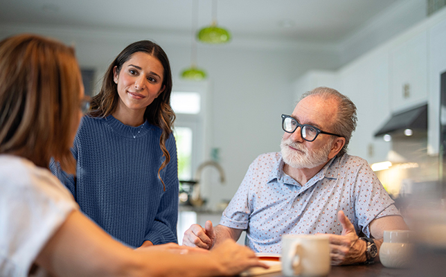 Three people seated around a table in a bright kitchen, reviewing documents and discussing information together.