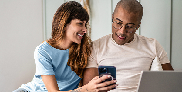 Woman in blue t-shirt and man in white t-shirt are looking at a mobile device. Man has a laptop on his lap