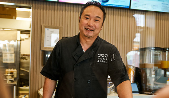 Restaurant employee in a black chef jacket standing in a food service area with menu displays and kitchen equipment behind them.