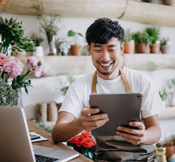 Man wearing an apron sitting at a desk in front of a laptop looking at a tablet device.