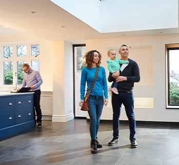 A woman is standing with a man holding a baby in an empty living room. A man is standing at a kitchen island looking at a binder