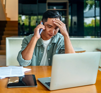 A man wearing a green shirt with his hand on his head is holding a mobile device to his ear while looking at a laptop