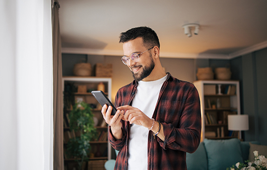 Man wearing glasses, white undershirt and plaid shirt is looking at mobile device in warm lit living room, standing in front of window.