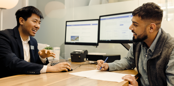 two men sitting at an office desk.  One man is holding a pen about to sign papers.