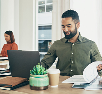 Man wearing green shirt at desk looking at laptop.  Woman in the background wearing red blouse.